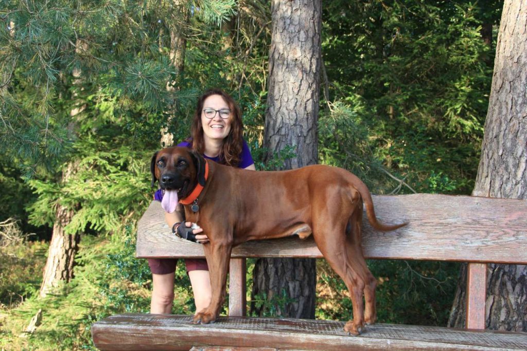 Katja mit ihrem Ridgeback Floyd auf einer Holzbank am Waldrand in der Lüneburger Heide – ein glücklicher Moment auf einem Spaziergang in der Natur der Nordheide.