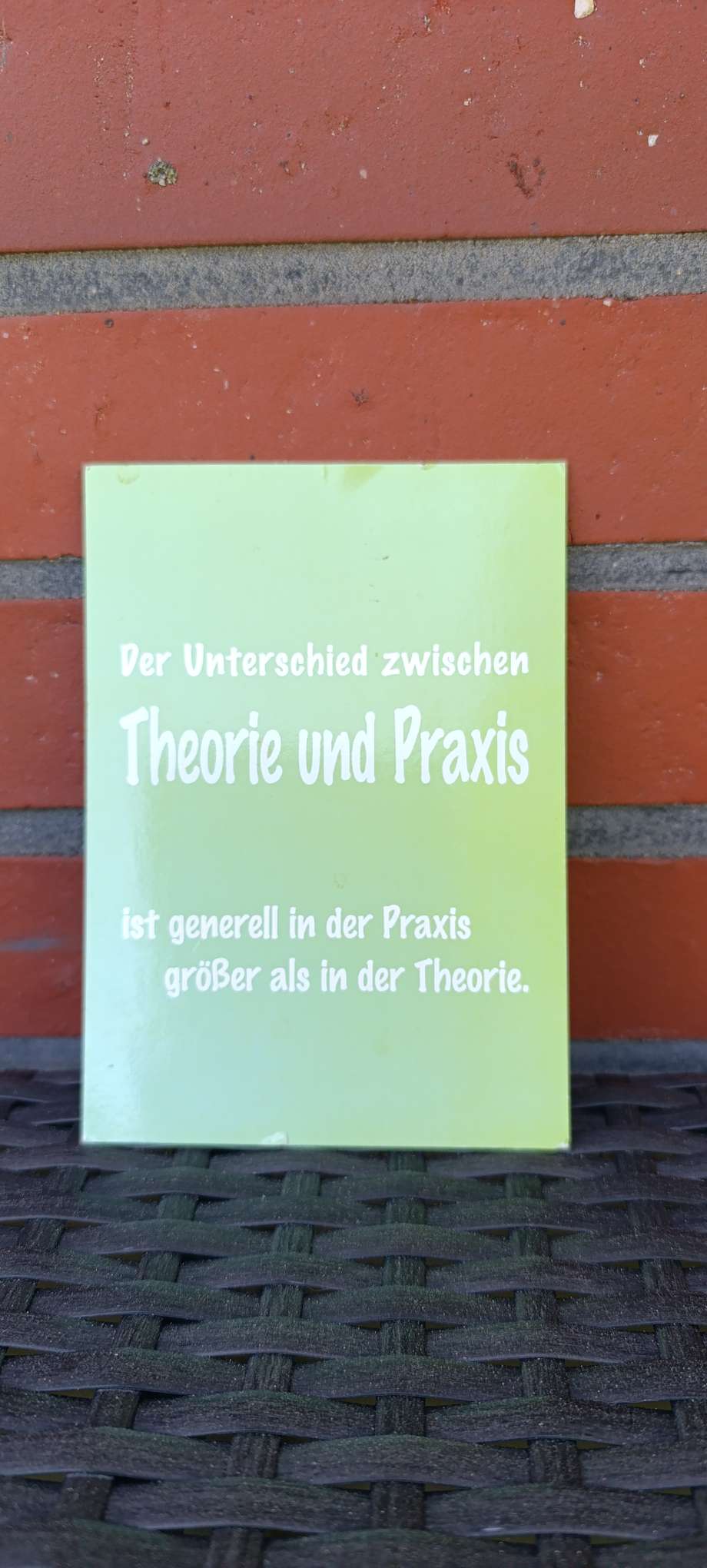 Postkarte mit dem Spruch ‚Der Unterschied zwischen Theorie und Praxis ist generell in der Praxis größer als in der Theorie‘ vor einer roten Backsteinwand – fotografiert als persönlicher Erinnerungsimpuls aus der Ausbildung zur Sozialpädagogischen Assistentin (SPA).