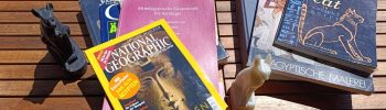 Bücher über Ägyptologie neben ägyptischen Katzenfiguren auf einem Gartentisch.
