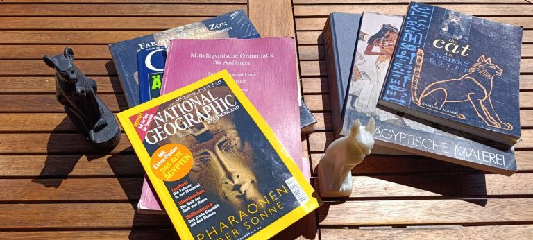 Bücher über Ägyptologie neben ägyptischen Katzenfiguren auf einem Gartentisch.