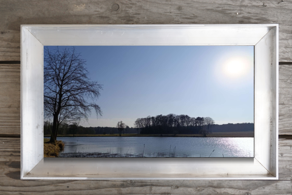 Gerahmtes Landschaftsbild mit See, Bäumen und Frühlingssonne – ruhiger Moment einer Schwellenzeit zwischen Winter und Frühling.