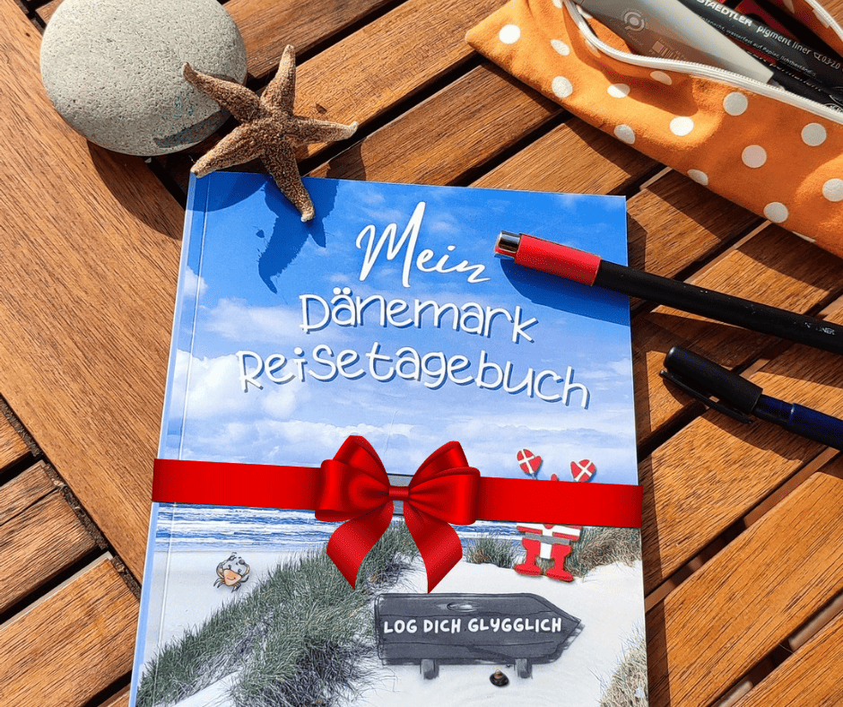 Mein Dänemark Reisetageuch als Geschenk mit rotem Schleifchen auf einem Gartentisch in der Sonne.
