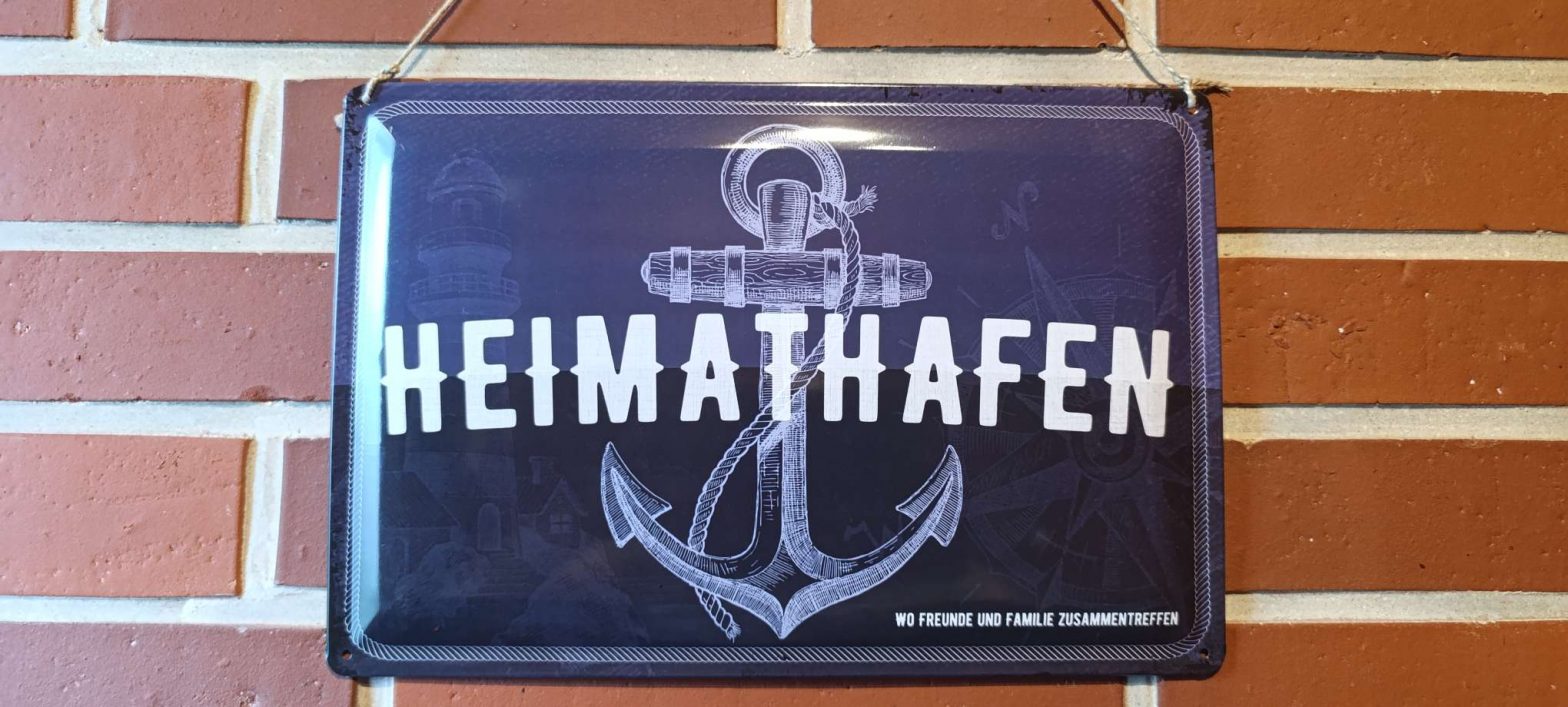 Blechschild mit der Aufschrift „Heimathafen“ und Anker an einer Backsteinwand.