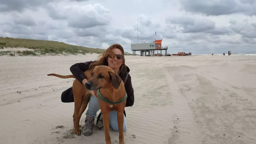 Katja kniet am windigen Strand und umarmt lächelnd ihre Rhodesian-Ridgeback-Hündin Keira