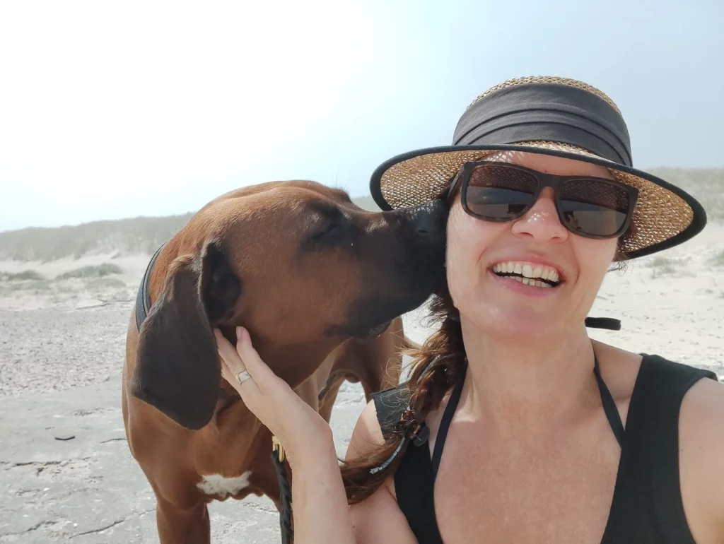 Rhodesian Ridgeback Floyd knabbert der lachenden Katja mit Sonnenhut am Strand am Ohr